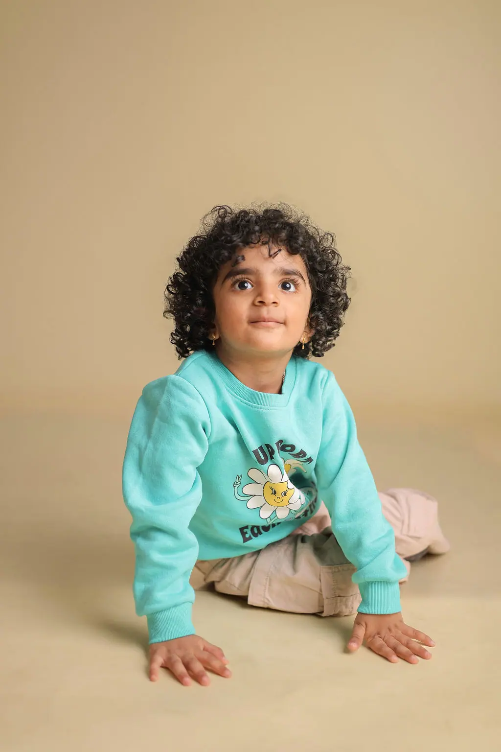Curly-haired toddler in a mint sweatshirt sitting on the floor and gazing up, plain beige backdrop, toddler photoshoot at WonderBump Studio, Bengaluru