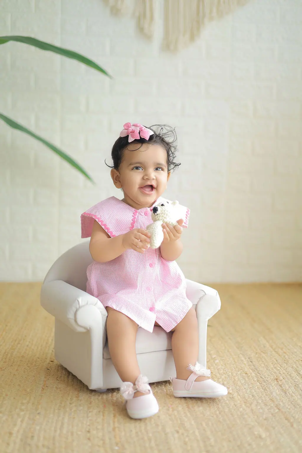 Laughing toddler in a pink gingham dress holding a crochet teddy on a small armchair, toddler photoshoot at WonderBump Studio, Bengaluru