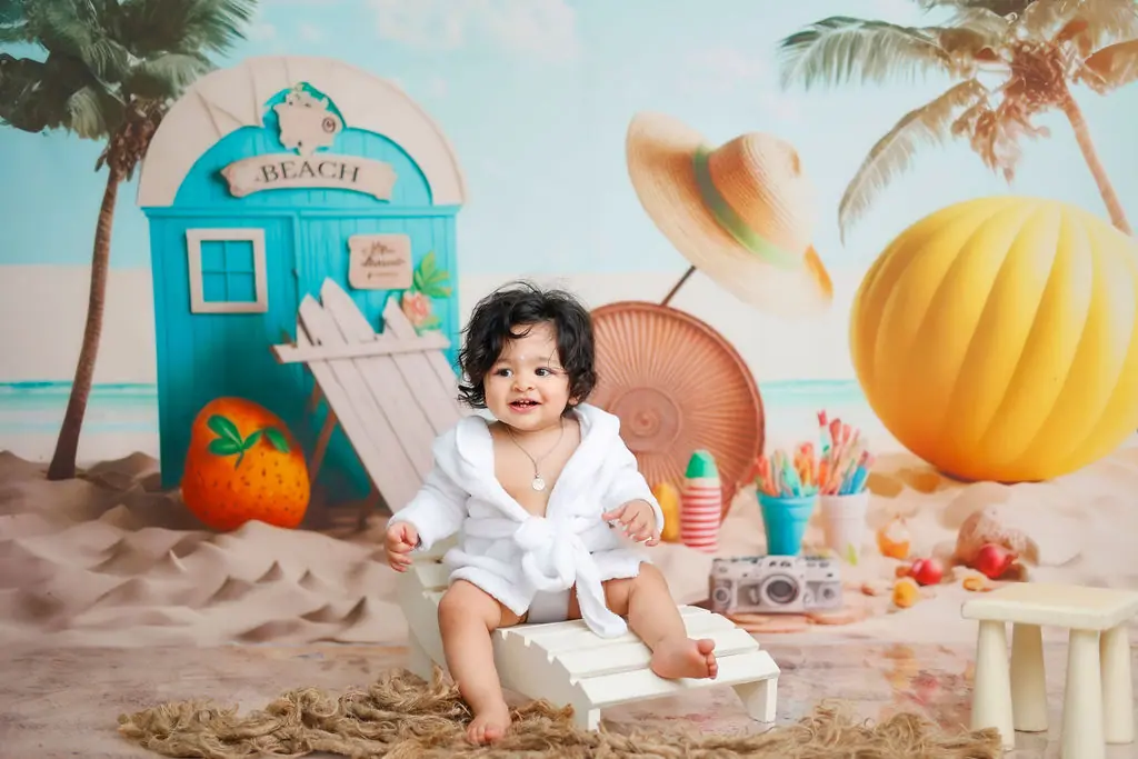 Smiling baby in a white bathrobe on a mini deckchair, printed beach backdrop, toddler photoshoot at WonderBump Studio, Bengaluru